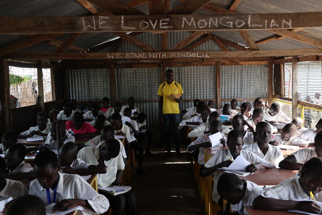 After 13 years of protecting civilians in Bentiu, the relationship between Mongolian peacekeepers and local communities is so strong that the children can even speak the Mongolian language. A classroom with students in white uniforms seated at wooden desks, writing in notebooks, while an instructor stands at the front under a corrugated metal roof with chalk writing above.