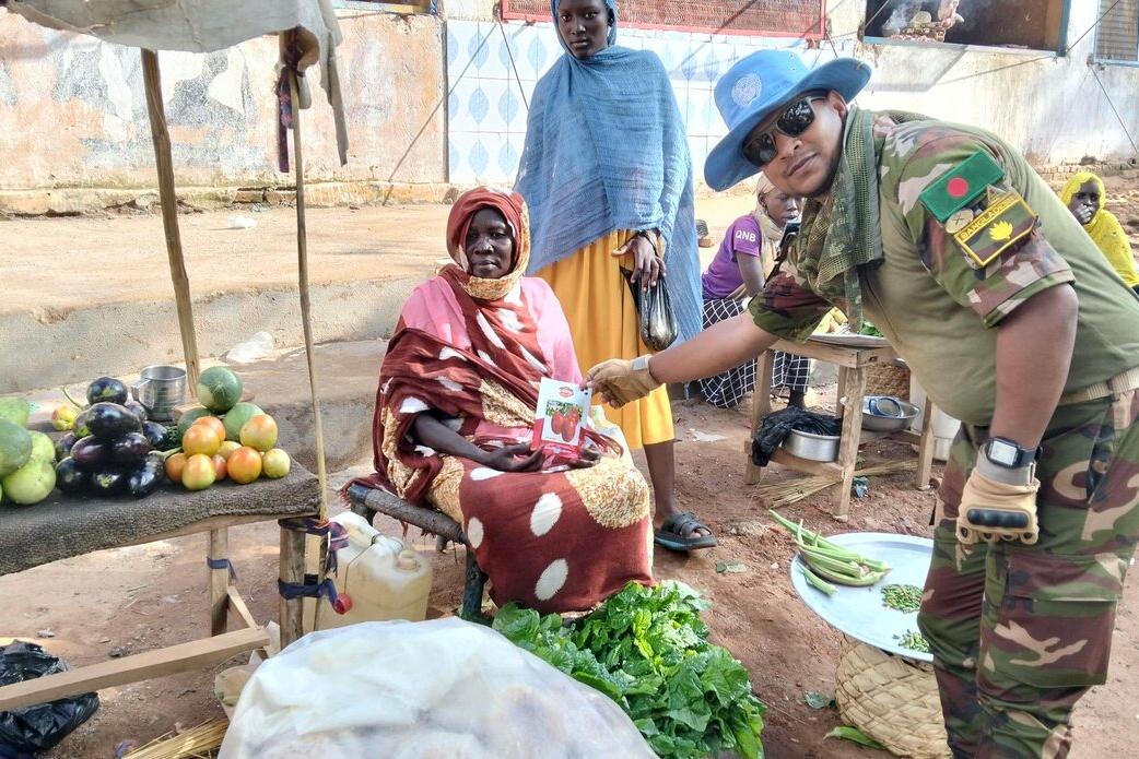 A person in camouflage uniform hands a leaflet to a seated vendor at an outdoor market stall with fruits and vegetables displayed on a mat