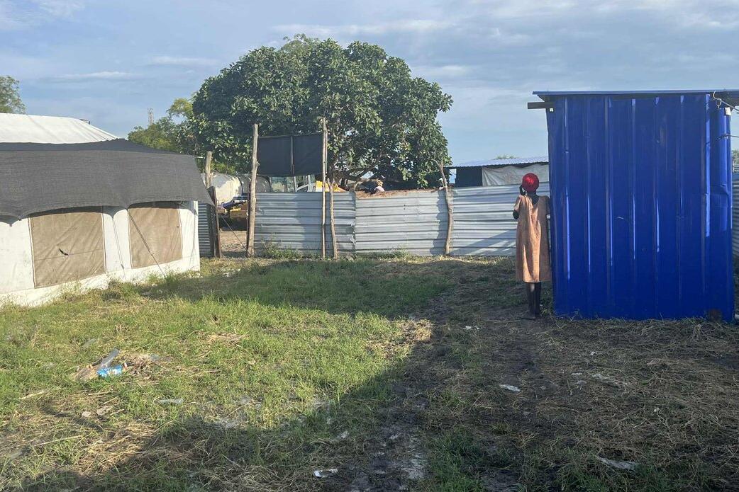 Outdoor area with grass and mud, showing a blue metal structure next to a fenced compound with tents and trees in the background. In the distance, a woman stands wearing an orange dress.