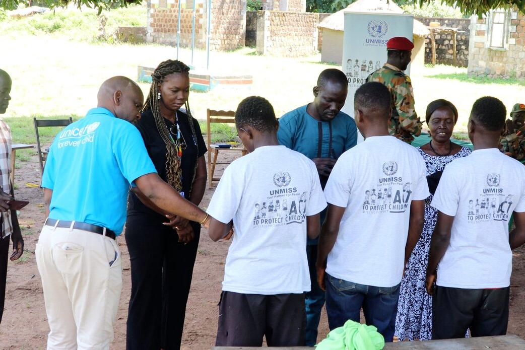 Group of people standing outdoors in a circle, including individuals wearing UNMISS-branded shirts and a person in a UNICEF shirt, with soldiers and chairs in the background.