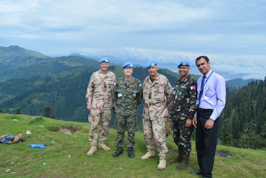 Four UN peacekeepers in blue berets and one official stand on a grassy hilltop with a scenic view of green mountains and cloudy skies in the background.